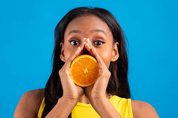 Young black woman holding orange slice in front of mouth on blue background