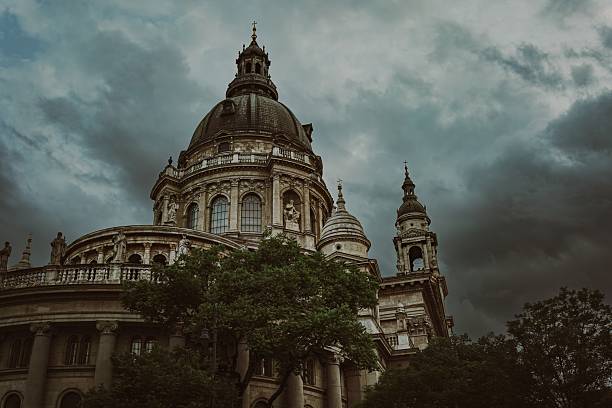 St. Stephen’s Basilica in Budapest, Hungary, captured from a low wide angle beneath turbulent storm clouds. The ornate statues, domes, and neoclassical details are framed by trees, creating a moody and cinematic scene rich in architectural grandeur and atmosphere.