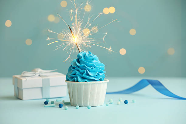 istock Birthday cupcake with burning sparkler and gift on light blue table against blurred lights 2224877805
