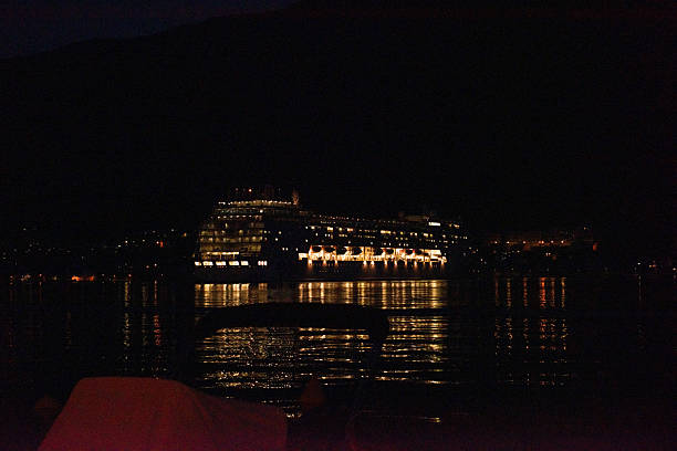 Kotor, Montenegro-05-06-2022: Large cruise ship sailing gracefully at night across the sea, with glowing windows casting reflections on the water, complemented by dark mountains rising in background