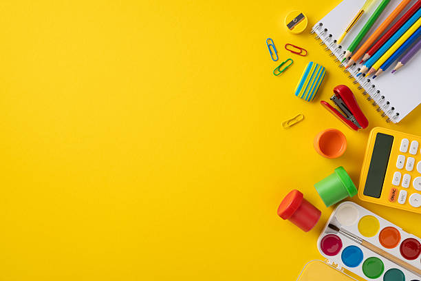 Educational items including pencils, notebook, watercolor paints, and a calculator arranged against a vivid yellow background, ideal for academic or creative themes