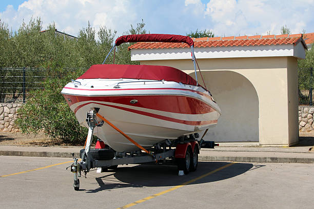 A sleek red and white fiberglass speedboat is tightly strapped to a double-axle trailer, parked in a sunlit lot beside a stucco shelter, ready for transport or storage
