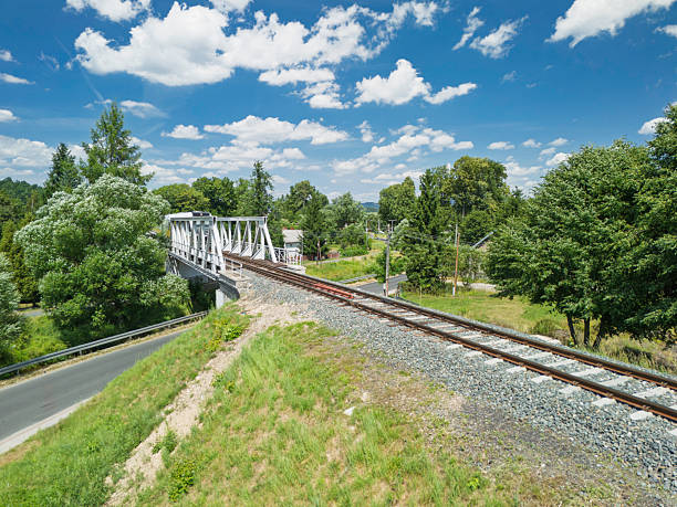 Track infrastructure spans across a bridge, facilitating rail travel in a rural setting. Lush trees surround the landscape, highlighting nature's beauty on a sunny day.