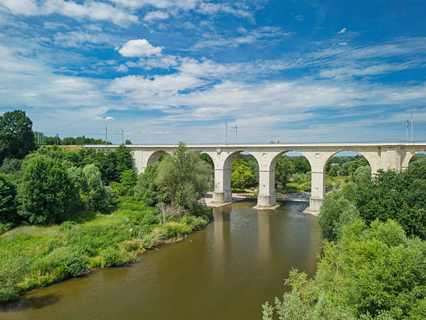 A well-structured railway viaduct crosses over a tranquil river, surrounded by vibrant greenery and blue skies, illustrating effective rail travel infrastructure on a clear day.