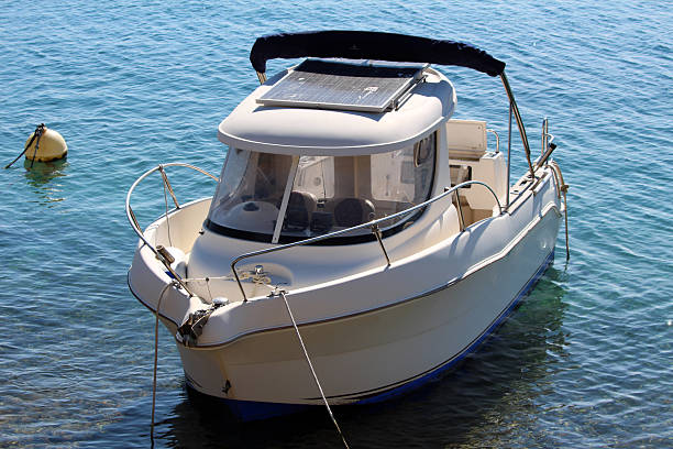 A compact white motorboat with a solar panel mounted on its roof floats on calm, clear blue water, tethered near a yellow buoy, showcasing sustainable boating technology