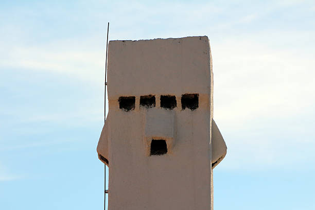 A white stucco chimney with five blackened vent openings and a vertical metal rod stands tall, its geometric design and weathered surface highlighted against a pale blue sky