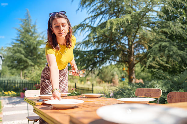 A young woman preparing a beautifully arranged table for an evening meal in the garden. The soft natural light and cozy atmosphere evoke a sense of warmth, hospitality, and seasonal outdoor living. Ideal for themes such as alfresco dining, home gatherings, slow living, and summer entertaining.