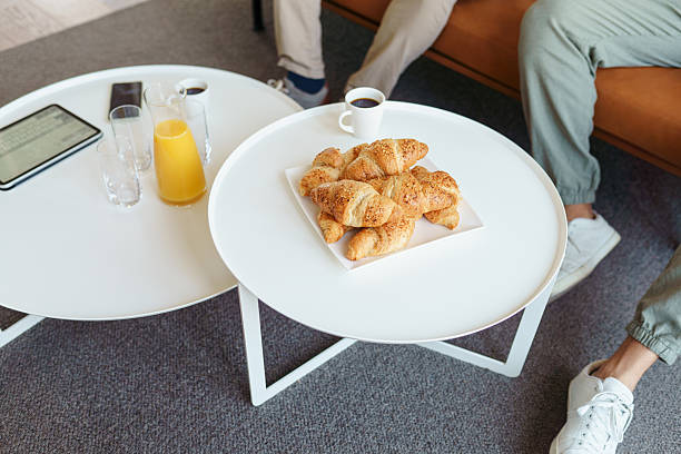 Group of colleagues relaxing around tables with croissants, juice, and coffee in an office lobby. Casual footwear and relaxed postures suggest a comfortable setting.
