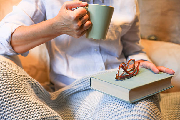 Senior Woman Enjoying A Relaxing Moment Indoors, Holding A Cup Of Hot Beverage While Resting A Closed Book With Glasses On Top. She Is Covered With A Soft Knit Blanket, Sitting Comfortably At Home, Creating A Cozy And Peaceful Atmosphere.