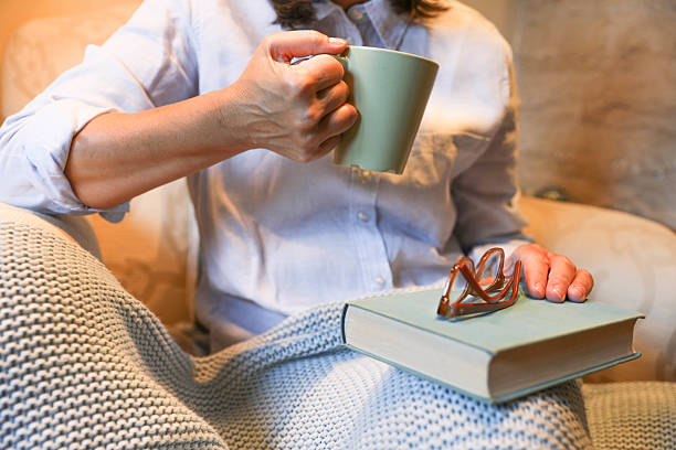 Senior Woman Enjoying A Relaxing Moment Indoors, Holding A Cup Of Hot Beverage While Resting A Closed Book With Glasses On Top. She Is Covered With A Soft Knit Blanket, Sitting Comfortably At Home, Creating A Cozy And Peaceful Atmosphere.