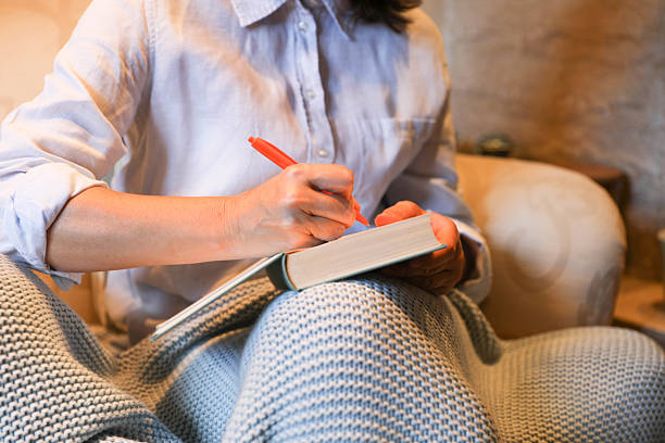 Senior Woman Sitting Comfortably Indoors, Covered With A Warm Knit Blanket, Reading And Taking Notes In A Book Using A Bright Colored Pen. The Scene Captures A Peaceful And Intellectual Moment Of Concentration And Learning In A Cozy Home Setting.