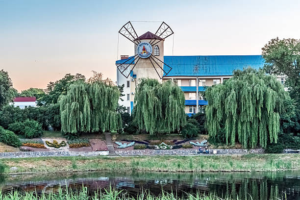 Historic windmill tower with blue roof dominates Myrhorod cityscape along Khorol River, surrounded by weeping willows and colorful flower gardens. Traditional Ukrainian landmark creates picturesque riverside scene with perfect water reflections. Myrhorod, Ukraine - June 29, 2024