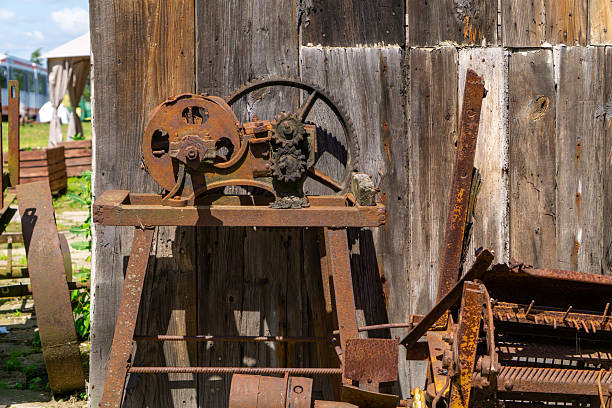 istock Rusty vintage mechanical saw with a belt-driven system, mounted on a wooden table in front of an old wooden shed. Visible gears and cast iron components suggest historical agricultural or workshop use. 2224928260