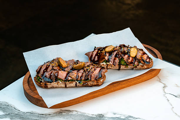 istock Gourmet bruschetta with pastrami, cream cheese, arugula, capers, and balsamic glaze on multigrain toast. Served on parchment over wooden tray. Perfect for restaurant menus, artisan cafes, catering promotions, and food styling projects. 2224929577