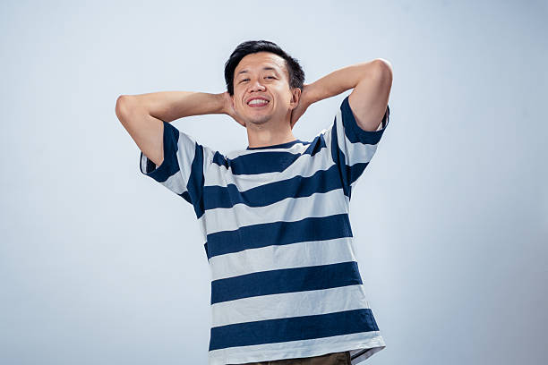 Asian man in a striped shirt smiling confidently with both hands placed behind his head, standing in front of a plain light background