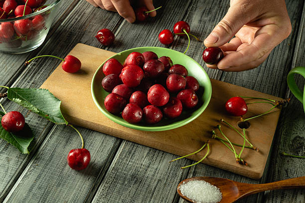 Hands are carefully selecting ripe cherries from a bowl in a rustic kitchen. Nearby, sugar and leaves create a natural ambiance, ready for a sweet creation