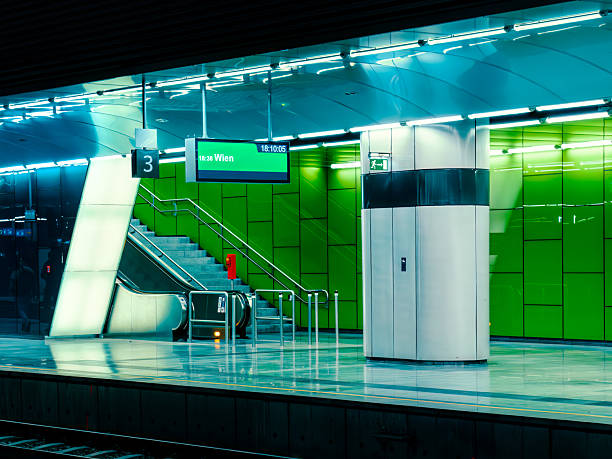 An interior view of a contemporary, brightly lit underground train station platform with vibrant green lighting and an escalator in the Vienna International Airport.