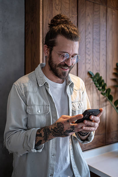 istock Smiling young man with tattoos and glasses using smartphone in modern kitchen 2224949651