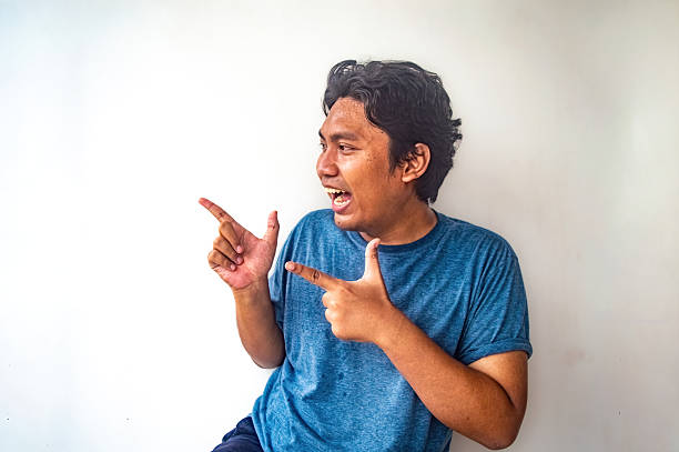 A man in a blue shirt is making a playful gesture with both hands, pointing in opposite directions. The plain white background makes the subject stand out clearly and captures an element of surprise