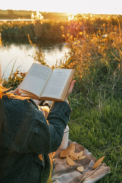 istock Girl by the river reading a book, beautiful sunset 2224998362