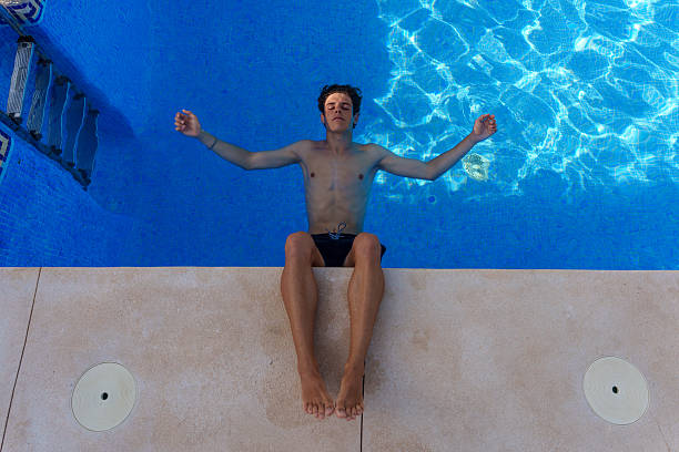 Young man relaxing floating in a blue swimming pool with open arms and eyes closed, enjoying summer holidays