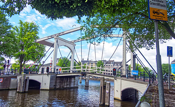 istock White drawbridge lifting over scenic amsterdam canal, flanked by verdant trees under vibrant azure sky 2225004410
