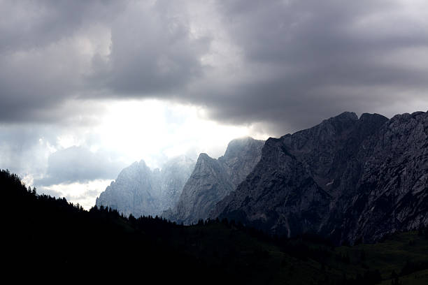 Towering mountains of Tyrol evoke a feeling of awe as dark clouds loom overhead, casting shadows and creating an ethereal atmosphere.