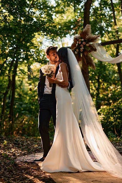 Young couple kissing in a lush forest during their wedding ceremony, celebrating their love amidst the beauty of nature, surrounded by sunlight filtering through the trees