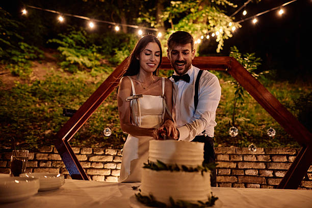 Bride and groom joyfully cutting their wedding cake together under twinkling string lights, celebrating their new life as husband and wife during an enchanting outdoor reception