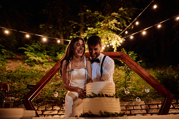 Happy newlyweds joyfully cutting their wedding cake together at an enchanting outdoor reception, illuminated by twinkling string lights, celebrating their love and lifelong commitment