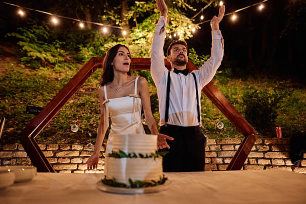 Surprised bride and groom raising their arms high to shield the wedding cake from falling decorations during an enchanting night reception filled with joy and celebration
