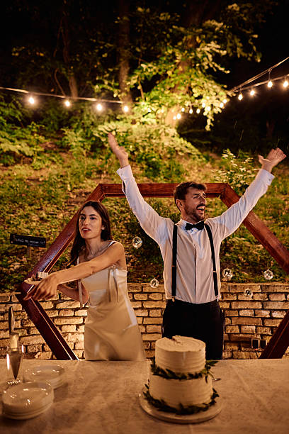 Bride and groom joyfully celebrating their wedding, raising their arms in excitement in front of their beautifully decorated cake during a magical outdoor evening party