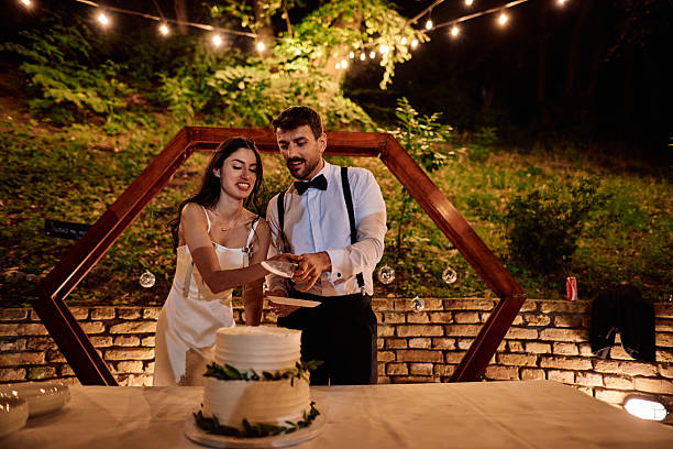 Newlyweds cutting their wedding cake together at an outdoor reception, celebrating their love and commitment under the romantic glow of string lights