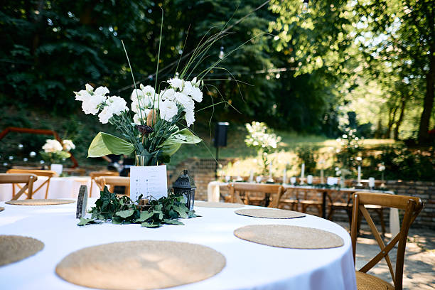 Round table with white tablecloth, centerpiece with white flowers and wicker placemats, wooden chairs and other tables in the background, all preparing for a wedding reception in a forest