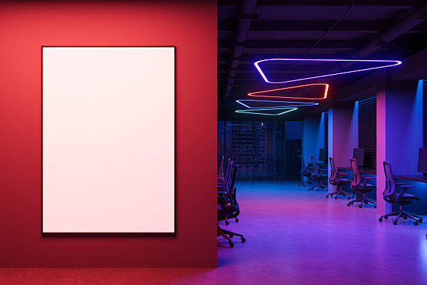 istock Blank vertical poster template on red wall in modern neon-lit office interior with computer desks and colorful ceiling lights at night. 3D Rendering 2225012386