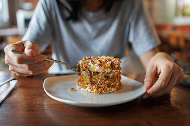 A woman in a grey shirt is seen holding a fork and plate, enjoying a slice of cake coated with chopped nuts at a cozy cafe.