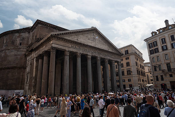 Large crowd of tourists exploring the historic Pantheon in Rome, showcasing its ancient Roman architecture under a partly cloudy sky. Overtourism concept.