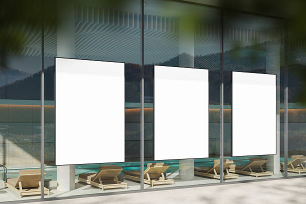 istock Three vertical blank poster mockups displayed on glass wall of modern building with minimalist poolside lounge chairs beneath them in natural daylight. 3D Rendering 2225019955