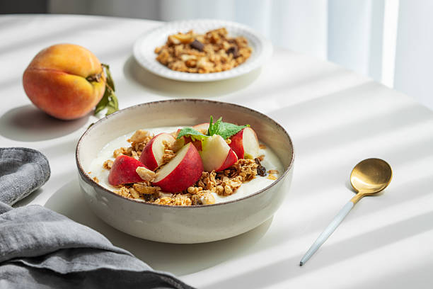 Natural greek yogurt with granola or muesli and peach in a bowl on a white table near window