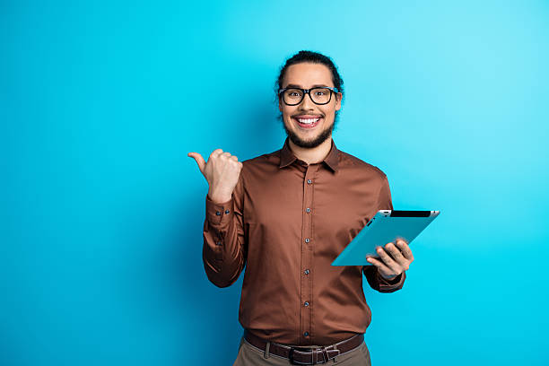 istock Confident young man pointing to the side while holding a tablet, standing against a bright turquoise background 2225035824