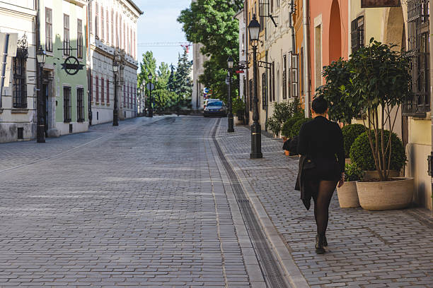 A woman dressed in black walks alone along a peaceful cobblestone street lined with pastel-colored historic buildings. The scene evokes calm, urban exploration, and timeless European charm.