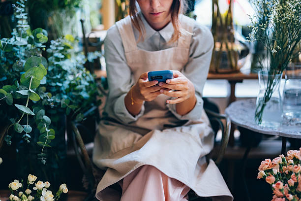 Florist Checking Smartphone While Surrounded by Blooming Flowers and Greenery