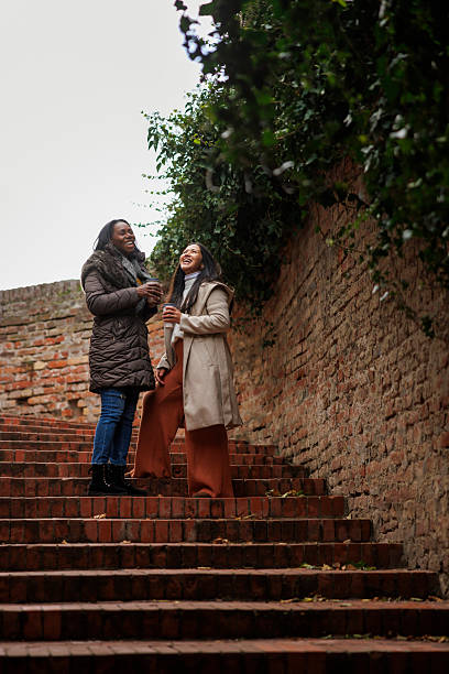 Two multi-ethnic female friends relaxing outdoors on cloudy autumn day, drinking coffee, chatting and taking a walk