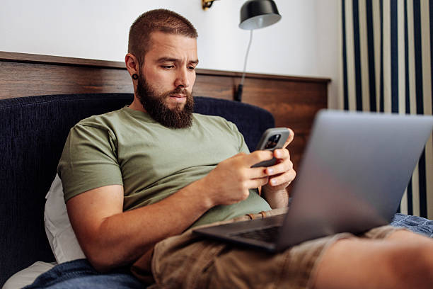 Man Using Smartphone and Laptop in Bedroom