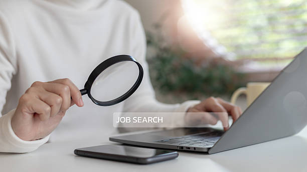 istock Person holding a magnifying glass while using a laptop with a job search text overlay. Concept of online job hunting, career search, employment opportunity, and recruitment. 2225072928