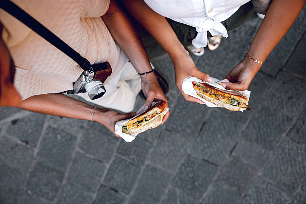 close-up shot of a couple enjoying local street food in Florence, Italy. They each hold a delicious sandwich, filled with fresh vegetables and savory ingredients, wrapped in paper for easy enjoyment. The woman, dressed in a white top and green pants, and the man, in a beige shirt, stand side by side, sharing a moment of culinary delight. The photo captures the simple joys of travel — exploring new flavors and savoring the experience together while immersed in the vibrant atmosphere of the city.