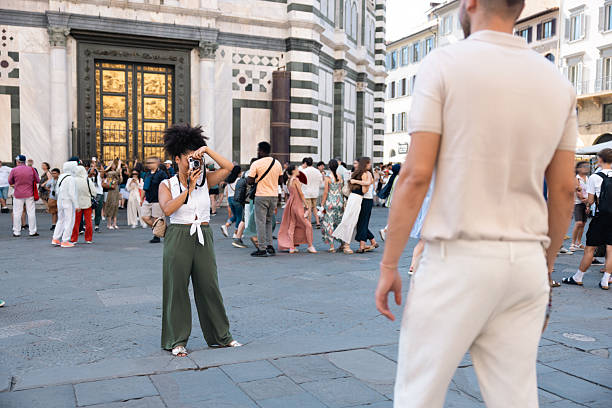 A woman stands in front of a grand building in Florence, Italy, taking a photo with her vintage camera. She’s dressed in a casual, summer outfit, with a white top and olive green wide-legged pants. In the background, a group of people walks by, some dressed in formal and casual summer clothes, adding to the lively atmosphere of the popular tourist destination. The soft, golden glow of the setting sun reflects off the historical architecture, adding a picturesque charm to the scene.