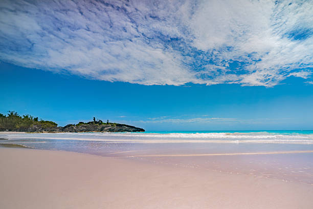 A pristine Horseshoe Bay Beach stretches out towards the horizon, featuring soft, pink sand meeting the gentle waves of turquoise waters. Fluffy clouds adorn the bright blue sky, while a small rock formation with greenery accentuates the scene in the background in Bermuda.