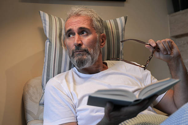 MATURE MAN With BEARD Relaxing On SOFA, Holding EYEGLASSES In HAND And BOOK In LAP. Thoughtful EXPRESSION Suggests TIREDNESS Or MOMENT OF PAUSE While READING. Concept Of AGING, VISION CARE, And HOME COMFORT.