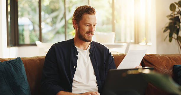 istock Man, paper and reading on sofa with laptop, writer and thinking for script draft in home. Person, editor and technology in living room for research, networking and application with freelancer 2225080160